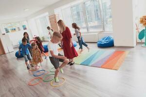 klein kinderkamer school- kinderen met vrouw leraar Aan verdieping binnenshuis in klas, aan het doen oefening. jumping over- hula hoepel cirkels bijhouden Aan de vloer. foto