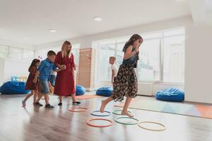 klein kinderkamer school- kinderen met vrouw leraar Aan verdieping binnenshuis in klas, aan het doen oefening. jumping over- hula hoepel cirkels bijhouden Aan de vloer. foto