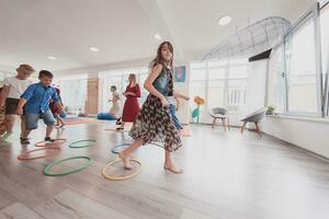 klein kinderkamer school- kinderen met vrouw leraar Aan verdieping binnenshuis in klas, aan het doen oefening. jumping over- hula hoepel cirkels bijhouden Aan de vloer. foto