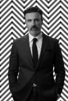zwart en wit portret van een elegant elegant senior zakenman met een baard en gewoontjes bedrijf kleren tegen retro kleurrijk patroon ontwerp achtergrond gebaren met handen foto