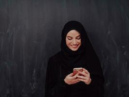 jong modern moslim bedrijf vrouw gebruik makend van smartphone vervelend hijab kleren in voorkant van zwart schoolbord foto