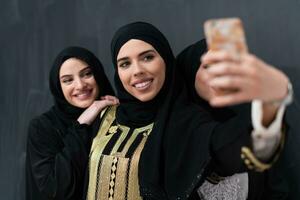 groep van jong mooi moslim Dames in modieus jurk met hijab gebruik makend van smartphone terwijl nemen selfie afbeelding in voorkant van zwart achtergrond foto