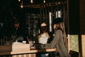 twee jong bedrijf Dames zittend Bij tafel in cafe. meisje shows collega informatie Aan laptop scherm. meisje gebruik makend van smartphone, bloggen. teamwerk, bedrijf ontmoeting.. foto