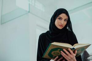 jong traditioneel moslim vrouw lezing koran Aan de sofa voordat iftar avondeten gedurende een Ramadan feest Bij huis foto