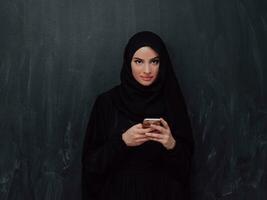 jong modern moslim bedrijf vrouw gebruik makend van smartphone vervelend hijab kleren in voorkant van zwart schoolbord foto