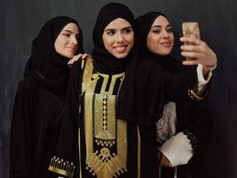 groep van jong mooi moslim Dames in modieus jurk met hijab gebruik makend van smartphone terwijl nemen selfie afbeelding in voorkant van zwart achtergrond foto