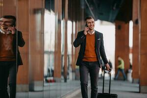 gaan naar luchthaven terminal. zelfverzekerd zakenman reiziger wandelen Aan stad straten en trekken zijn koffer drinken koffie en sprekend Aan smartphone foto