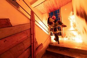 dapper brandweerman daalt af trap van een brandend gebouw en houdt opgeslagen meisje in zijn armen. Open brand en een brandweerman in de achtergrond. foto