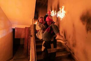 dapper brandweerman daalt af trap van een brandend gebouw en houdt opgeslagen meisje in zijn armen. Open brand en een brandweerman in de achtergrond. foto