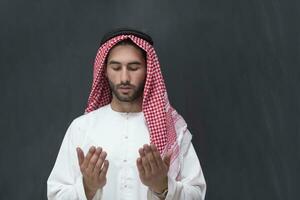 een jong Arabisch Mens in traditioneel kleren maken een traditioneel gebed naar god houdt zijn handen in bidden gebaar in voorkant van een zwart achtergrond foto