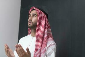 een jong Arabisch Mens in traditioneel kleren maken een traditioneel gebed naar god houdt zijn handen in bidden gebaar in voorkant van een zwart achtergrond foto