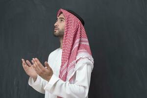 een jong Arabisch Mens in traditioneel kleren maken een traditioneel gebed naar god houdt zijn handen in bidden gebaar in voorkant van een zwart achtergrond foto