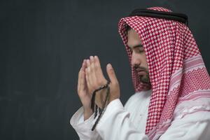 een jong Arabisch Mens in traditioneel kleren maken een traditioneel gebed naar god houdt zijn handen in bidden gebaar in voorkant van een zwart achtergrond foto