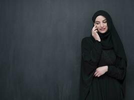 jong modern moslim bedrijf vrouw gebruik makend van smartphone vervelend hijab kleren in voorkant van zwart schoolbord foto