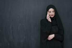 jong modern moslim bedrijf vrouw gebruik makend van smartphone vervelend hijab kleren in voorkant van zwart schoolbord foto