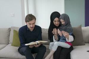 traditioneel moslim familie ouders met kinderen lezing koran en bidden samen Aan de sofa voordat iftar avondeten gedurende een Ramadan feest Bij huis foto