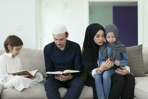 traditioneel moslim familie ouders met kinderen lezing koran en bidden samen Aan de sofa voordat iftar avondeten gedurende een Ramadan feest Bij huis foto