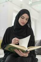 jong traditioneel moslim vrouw lezing koran Aan de sofa voordat iftar avondeten gedurende een Ramadan feest Bij huis foto