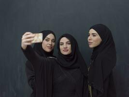 groep van jong moslim Dames in modieus jurk met hijab gebruik makend van smartphone terwijl nemen selfie afbeelding in voorkant van zwart achtergrond foto