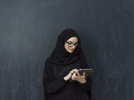 jong modern moslim bedrijf vrouw gebruik makend van smartphone vervelend zonnebril en hijab kleren in voorkant van zwart schoolbord foto