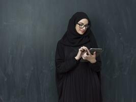 jong modern moslim bedrijf vrouw gebruik makend van smartphone vervelend zonnebril en hijab kleren in voorkant van zwart schoolbord foto