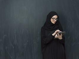 jong modern moslim bedrijf vrouw gebruik makend van smartphone vervelend zonnebril en hijab kleren in voorkant van zwart schoolbord foto