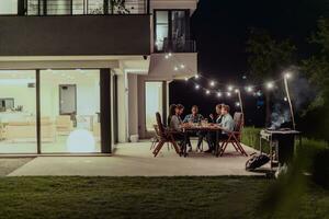 een groep van jong verschillend mensen hebben avondeten Aan de terras van een modern huis in de avond. pret voor vrienden en familie. viering van vakantie, bruiloften met barbecue. foto