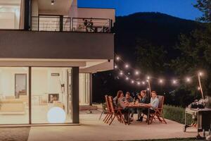 een groep van jong verschillend mensen hebben avondeten Aan de terras van een modern huis in de avond. pret voor vrienden en familie. viering van vakantie, bruiloften met barbecue. foto