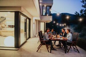 een groep van jong verschillend mensen hebben avondeten Aan de terras van een modern huis in de avond. pret voor vrienden en familie. viering van vakantie, bruiloften met barbecue. foto