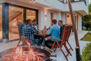 een groep van jong verschillend mensen hebben avondeten Aan de terras van een modern huis in de avond. pret voor vrienden en familie. viering van vakantie, bruiloften met barbecue. foto