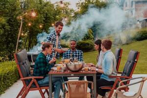 een groep van jong verschillend mensen hebben avondeten Aan de terras van een modern huis in de avond. pret voor vrienden en familie. viering van vakantie, bruiloften met barbecue. foto