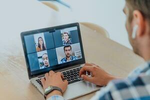 de Mens zittend Bij een tafel in een modern leven kamer, met hoofdtelefoons gebruik makend van een laptop voor bedrijf video chatten, gesprek met vrienden en vermaak foto