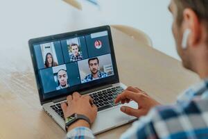 de Mens zittend Bij een tafel in een modern leven kamer, met hoofdtelefoons gebruik makend van een laptop voor bedrijf video chatten, gesprek met vrienden en vermaak foto