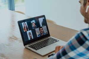 de Mens zittend Bij een tafel in een modern leven kamer, met hoofdtelefoons gebruik makend van een laptop voor bedrijf video chatten, gesprek met vrienden en vermaak foto