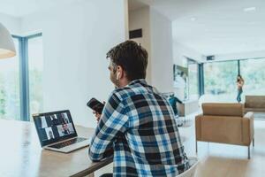 de Mens zittend Bij een tafel in een modern leven kamer, gebruik makend van een smartphone en laptop voor bedrijf video chatten, gesprek met vrienden en vermaak foto