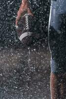 dichtbij omhoog van Amerikaans Amerikaans voetbal atleet krijger staand Aan een veld- focus Aan bal en klaar naar Speel. speler voorbereidingen treffen naar rennen, aanval en partituur touchdown. regenachtig nacht met dramatisch lens gloed en regen druppels. foto