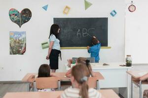 elementair school. de vrouw leraar helpen de kind leerling terwijl schrijven de antwoord Aan de schoolbord. foto