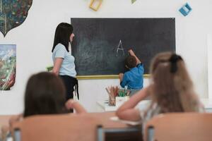 elementair school. de vrouw leraar helpen de kind leerling terwijl schrijven de antwoord Aan de schoolbord. foto