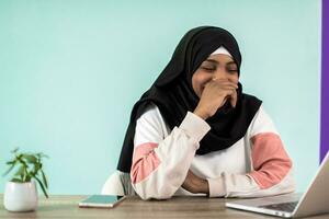 Afrikaanse Amerikaans vrouw vervelend een hijab bedachtzaam zittend in haar huis kantoor en toepassingen een laptop en smartphone foto