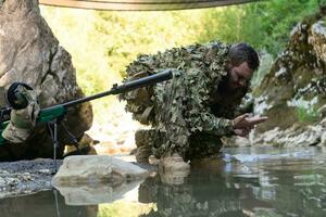 soldaat in een camouflage pak uniform drinken vers water van de rivier. leger scherpschutter geweer- Aan de kant. foto