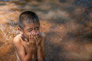 Aziatisch jong jongen schattig en charmant het wassen gezicht in rivier- Aan achtergrond water oppervlakte en zonsopgangen, kind spelen water, levensstijl van Aziatisch kinderen, begrip van leven en duurzaamheid. foto