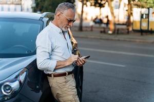 man van middelbare leeftijd die in de buurt van zijn suv-auto staat en zijn smartphone gebruikt. foto