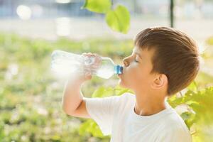 schattig jongen zittend Aan de gras drankjes water van een fles in de zomer Bij zonsondergang. kind blust dorst Aan een heet dag foto