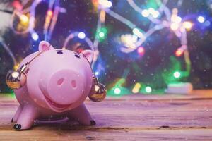 opslaan geld voor kerstmis. varkentje bank met Kerstmis speelgoed Aan oren tegen versierd met feestelijk lichten Kerstmis boom. Kerstmis financiën concept. foto