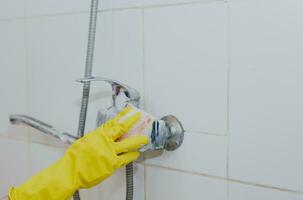 huisvrouw schoonmaak badkamer kraan en douche tik. hulp in de huishouding in geel beschermend handschoenen het wassen vuil bad tik. handen van vrouw het wassen bad foto