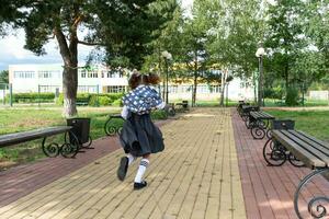 vrolijk grappig meisje met tandeloos glimlach in school- uniform met wit bogen rennen in school- tuin. terug naar school, september 1. gelukkig leerling met rugzak. primair opleiding, elementair klas. beweging foto