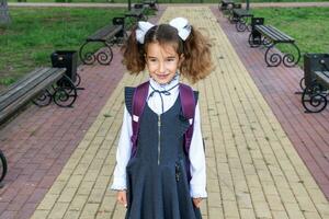 vrolijk grappig meisje met een tandeloos glimlach in een school- uniform met wit bogen in school- tuin. terug naar school, september 1. gelukkig leerling met een rugzak. primair opleiding, elementair klas. foto