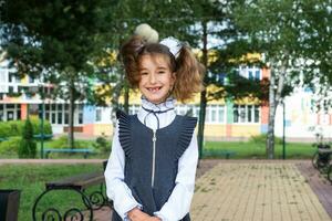 vrolijk grappig meisje met een tandeloos glimlach in een school- uniform met wit bogen in school- tuin. terug naar school, september 1. een gelukkig leerling. primair opleiding, elementair klas. portret van een leerling foto