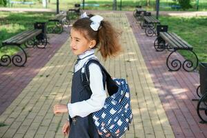 vrolijk grappig meisje met een tandeloos glimlach in een school- uniform met wit bogen in school- tuin. terug naar school, september 1. gelukkig leerling met een rugzak. primair opleiding, elementair klas. foto