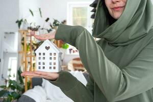 een gelukkig vrouw in haar huis houdt in haar handen een miniatuur figuur van een huis en sleutel in de interieur. droom huis projecteren, echt landgoed aankoop, verzekering, hypotheek, huur, reservering foto